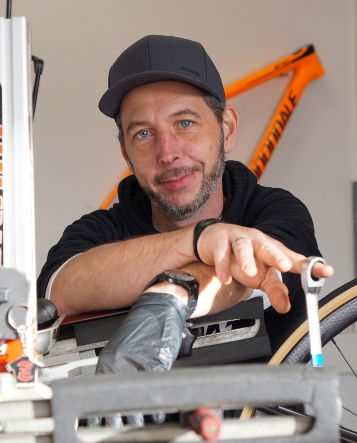 Mechaniker mit Basecap beim Zentrieren eines Laufrads in der Werkstatt – orangefarbenes Fahrradrahmen im Hintergrund.